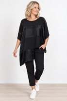 A woman modeling a black asymmetrical short-sleeve top with textured paneling, paired with black pants and white sneakers, against a clean white studio background.
