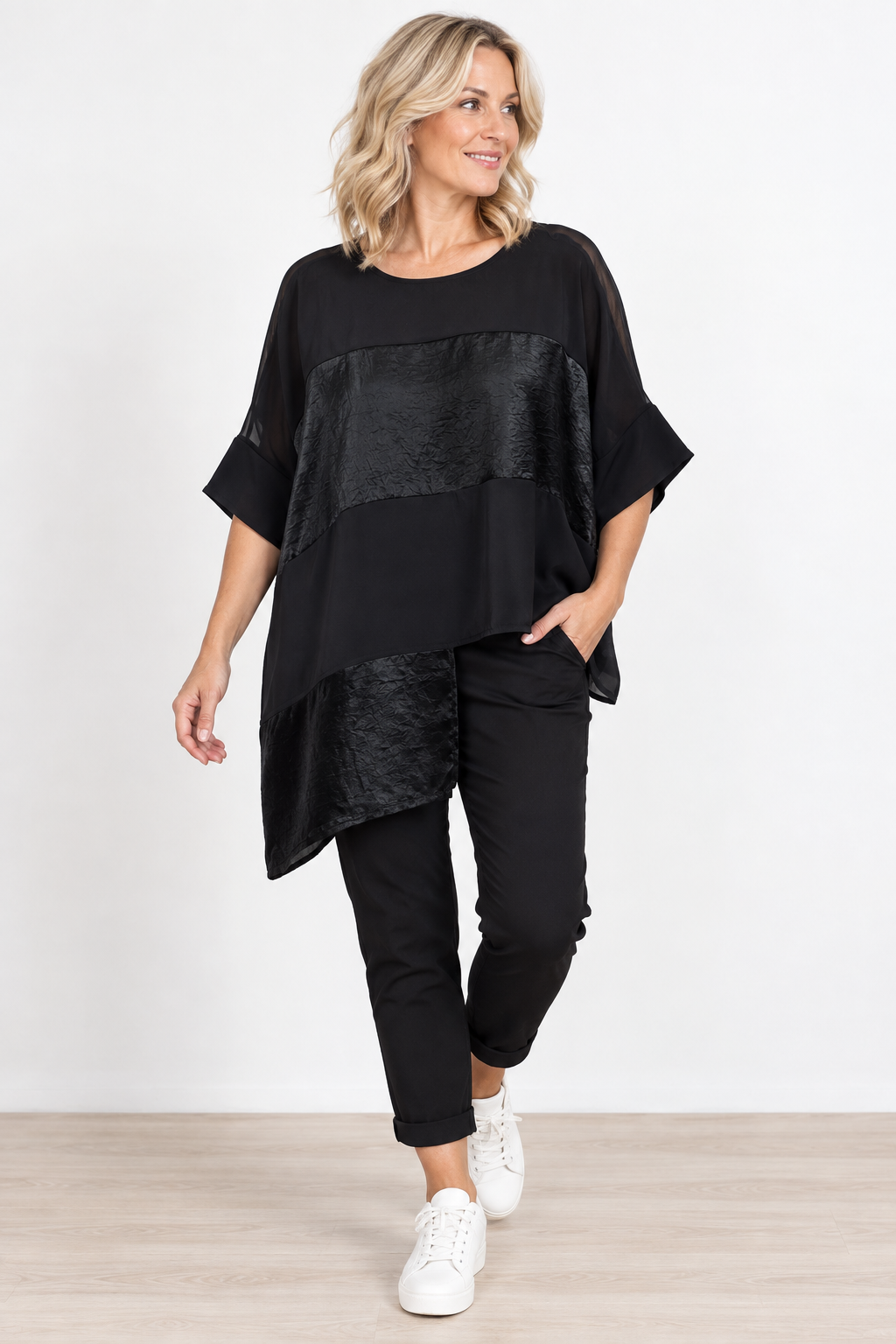 A woman modeling a black two-piece outfit consisting of a short-sleeve, asymmetrical top with textured paneling and matching black cropped pants, paired with white sneakers, against a plain white studio background.