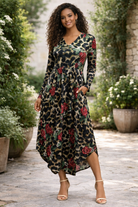 Woman wearing a long floral dress in an outdoor setting with plants and stone pavement.