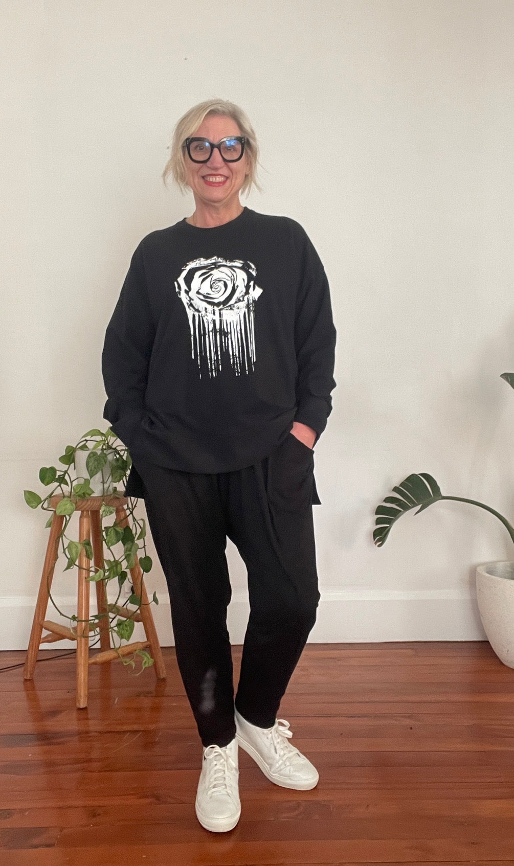 Person wearing a black sweatshirt with a white graphic design, black pants, and white sneakers in a room with plants and a vase.