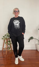 Person wearing a black sweatshirt with a white graphic design, black pants, and white sneakers in a room with plants and a vase.