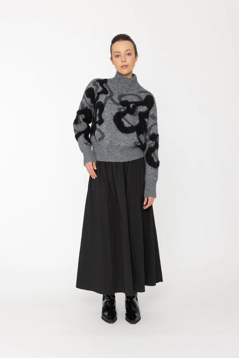Person wearing a gray sweater with black abstract patterns and a long black skirt on a white background