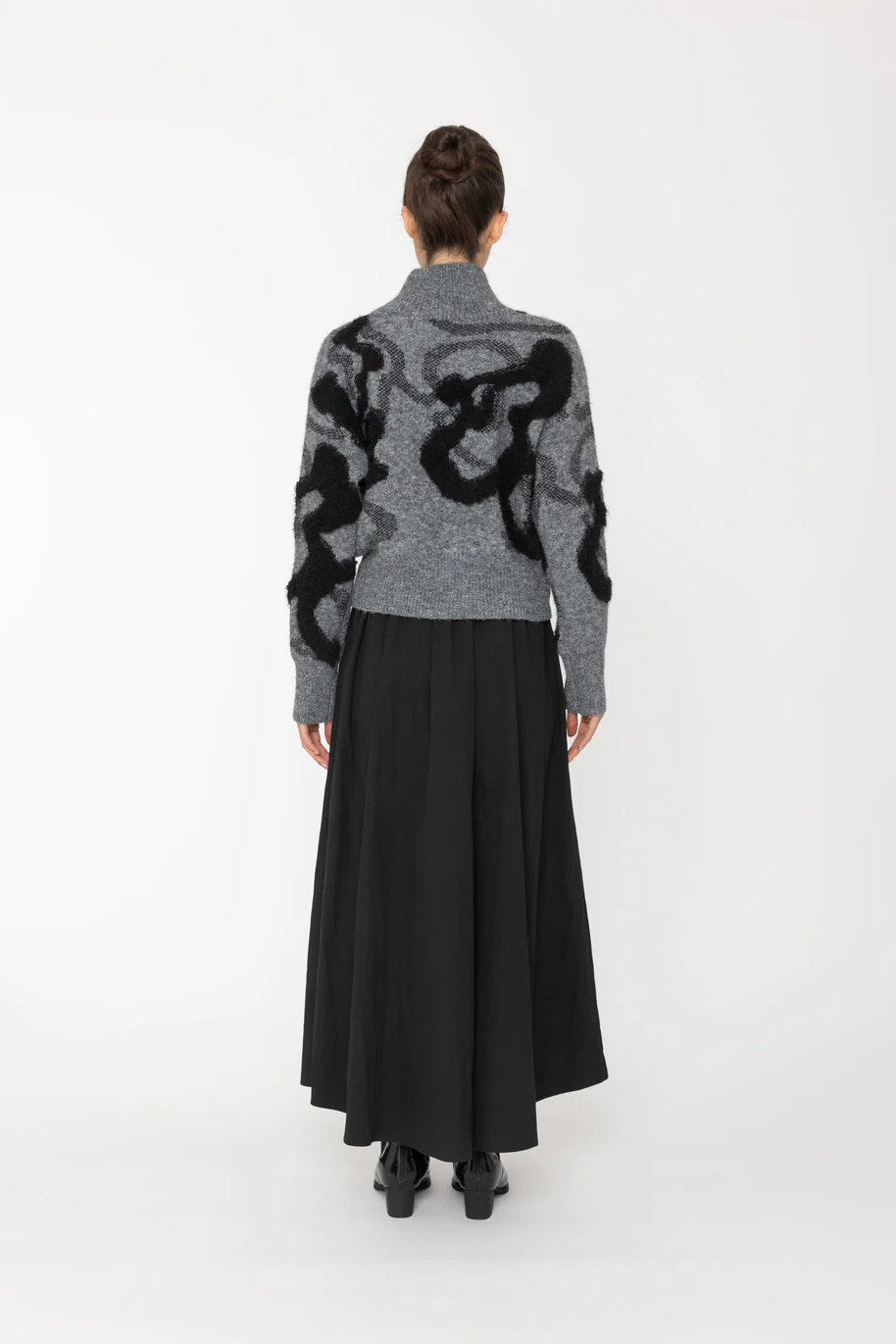 Person wearing a gray sweater with black abstract patterns and a long black skirt on a white background