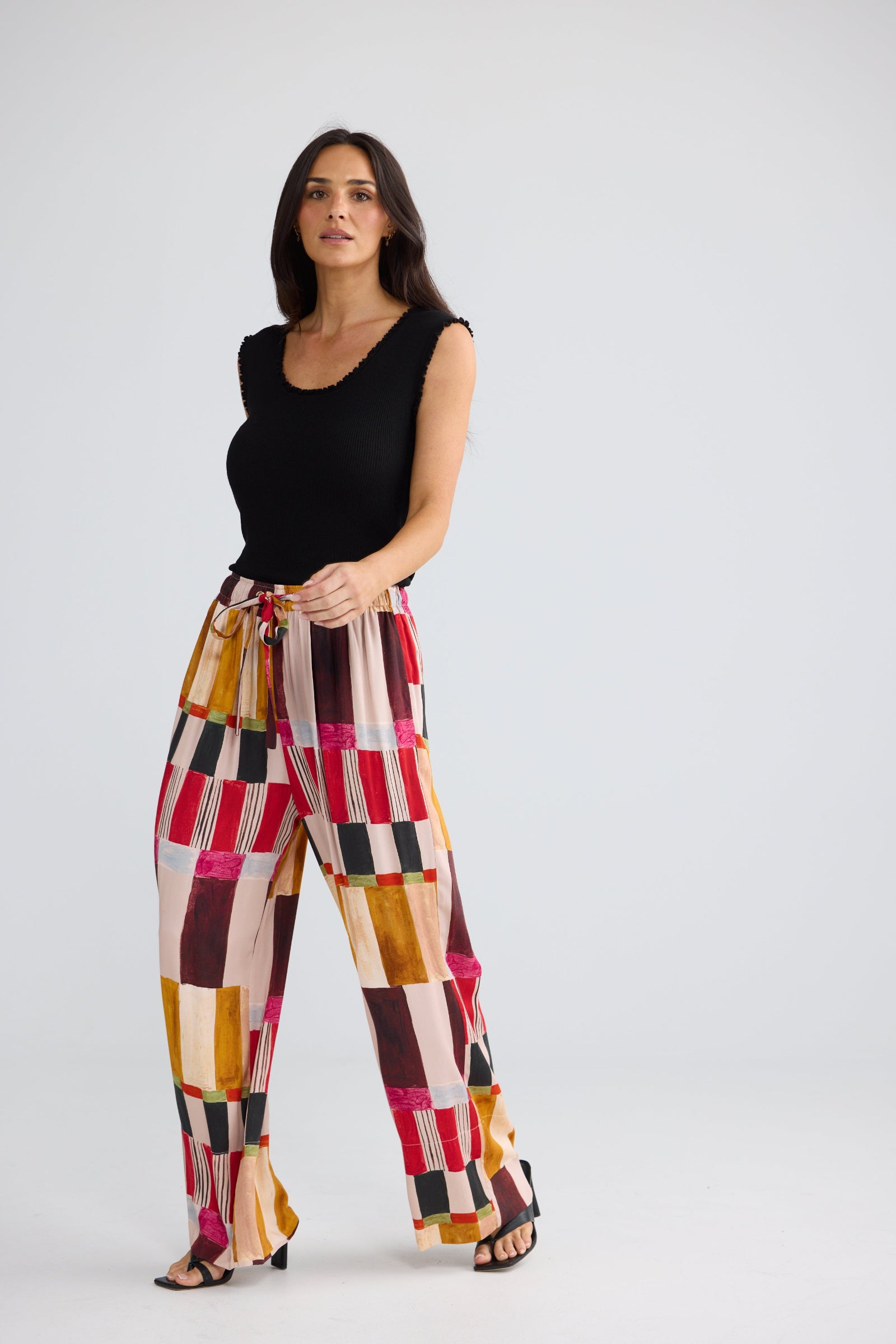 A woman modeling high-waisted, wide-leg trousers with a colorful geometric patchwork pattern in pink, orange, yellow, red, and black, paired with a black sleeveless top, against a light gray studio background.