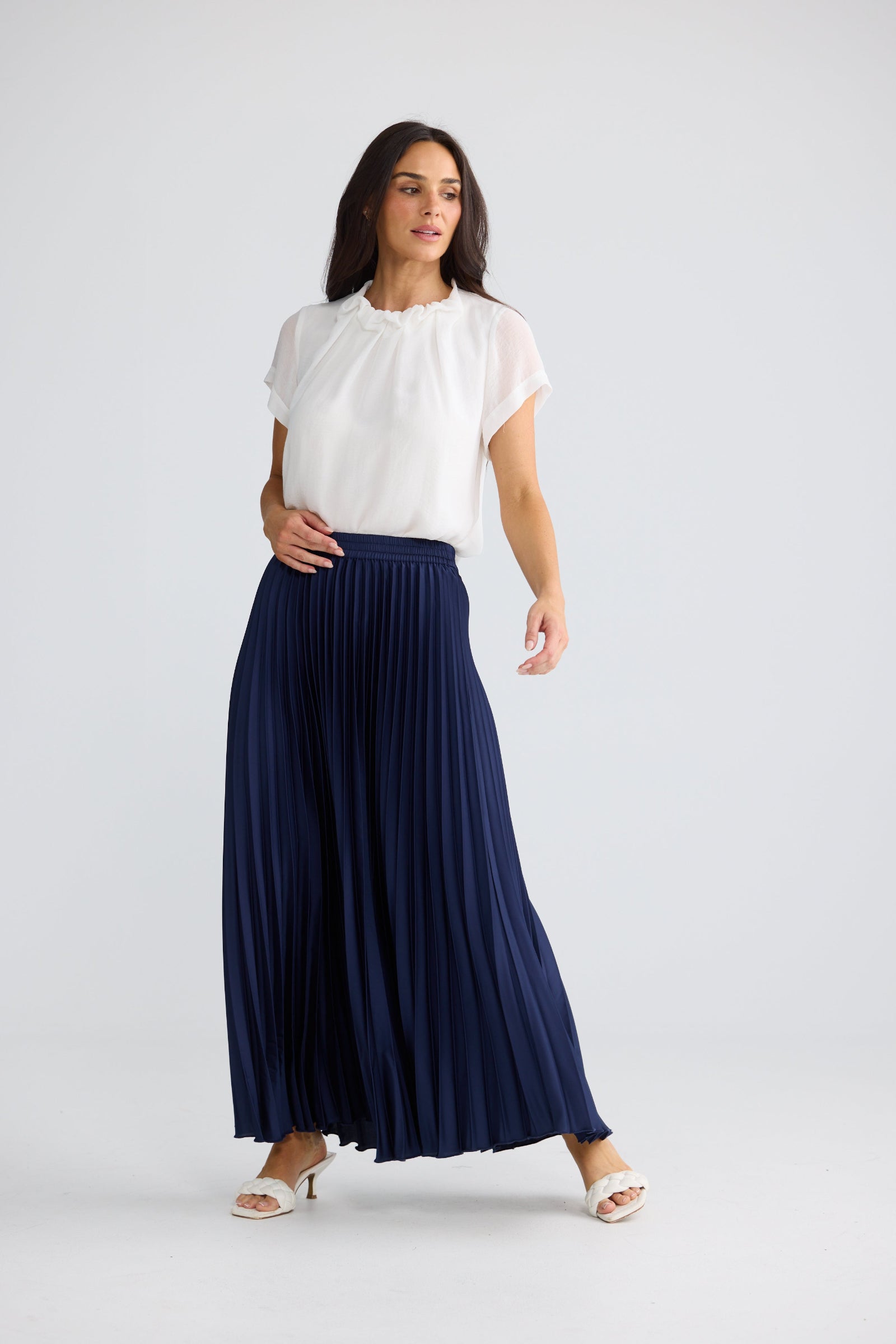 A woman modeling a white short-sleeve blouse and a navy blue pleated maxi skirt against a light gray studio background.