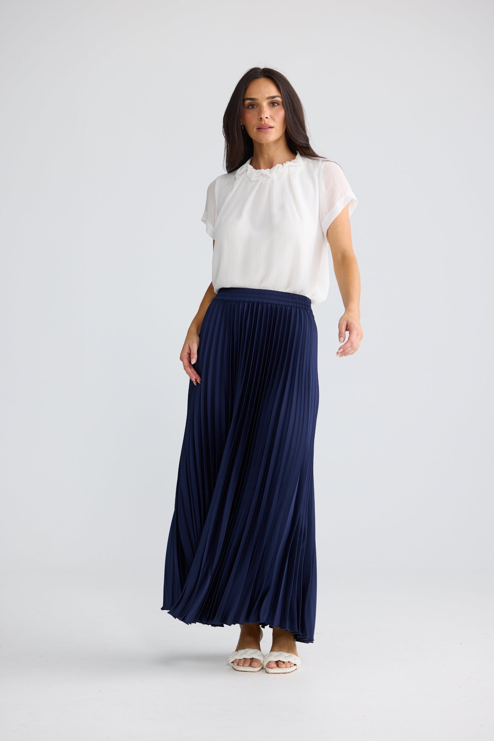 A woman modeling a white short-sleeve blouse and a navy blue pleated maxi skirt, standing against a light gray studio background.