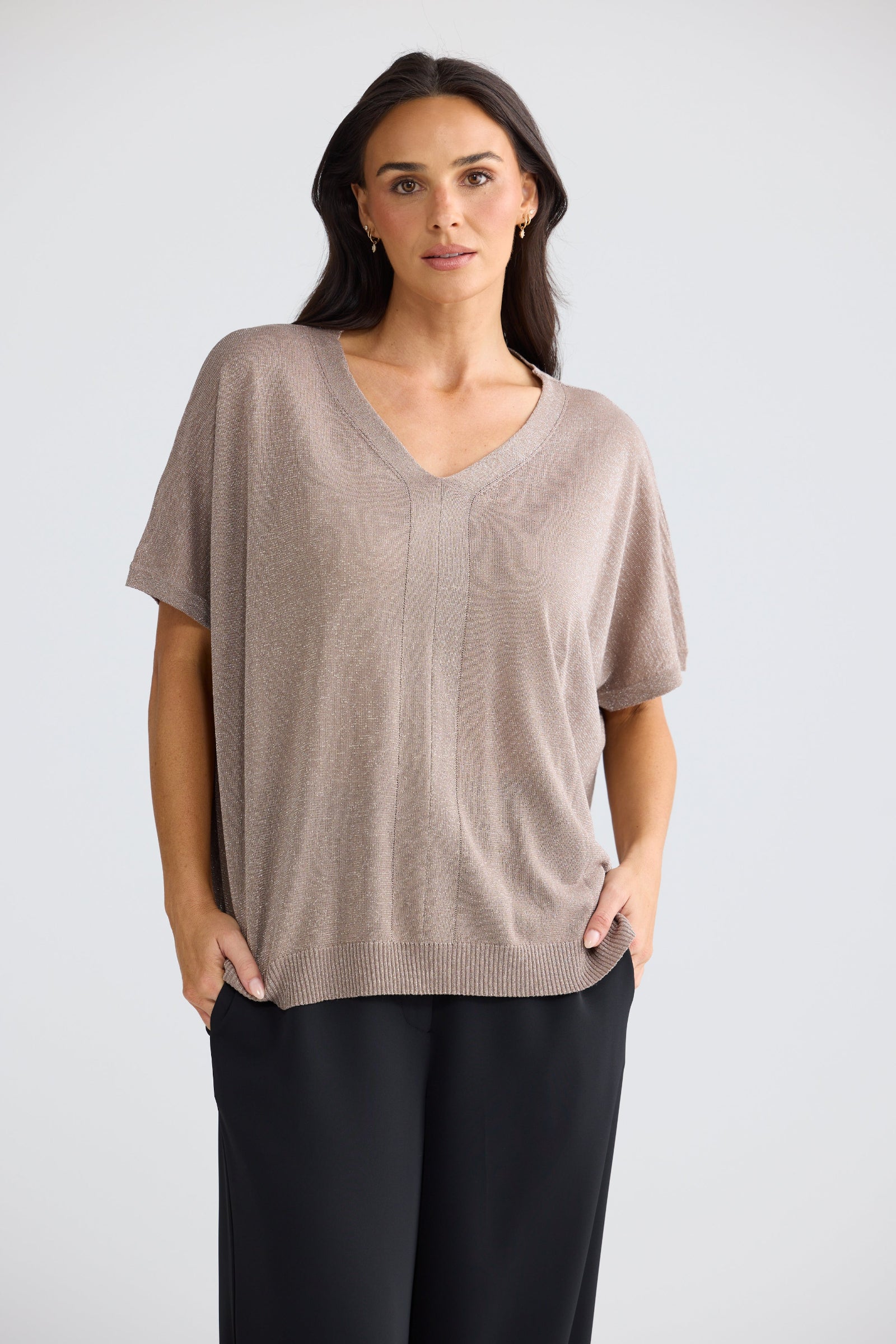 A woman modeling a short-sleeve knit V-neck top in a light taupe-beige color, paired with black pants, against a plain light gray studio background.