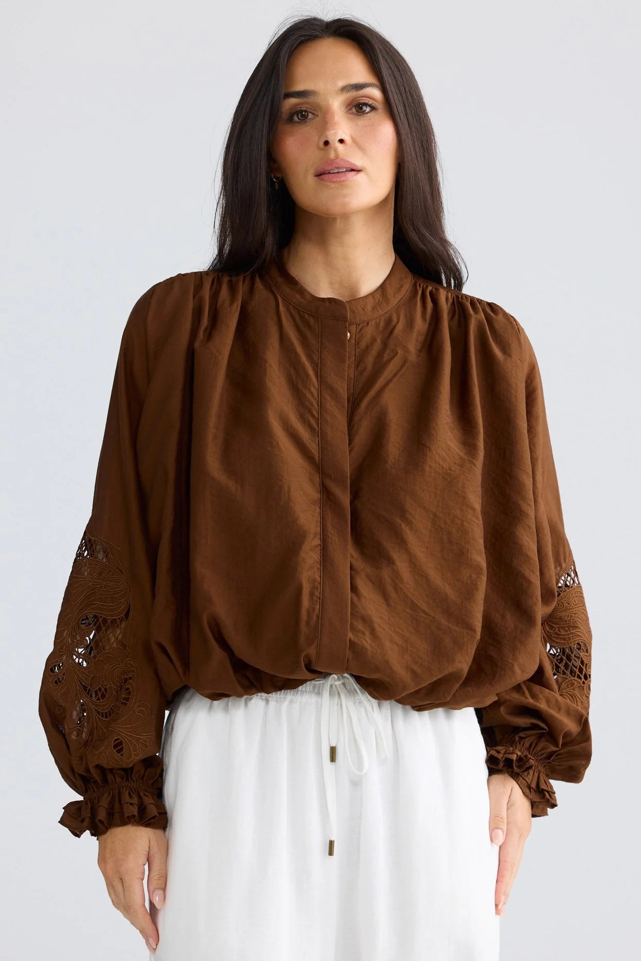 A woman wearing a brown long-sleeve blouse with gathered, balloon sleeves and embroidered details on the cuffs, paired with white drawstring pants, against a light gray studio background.