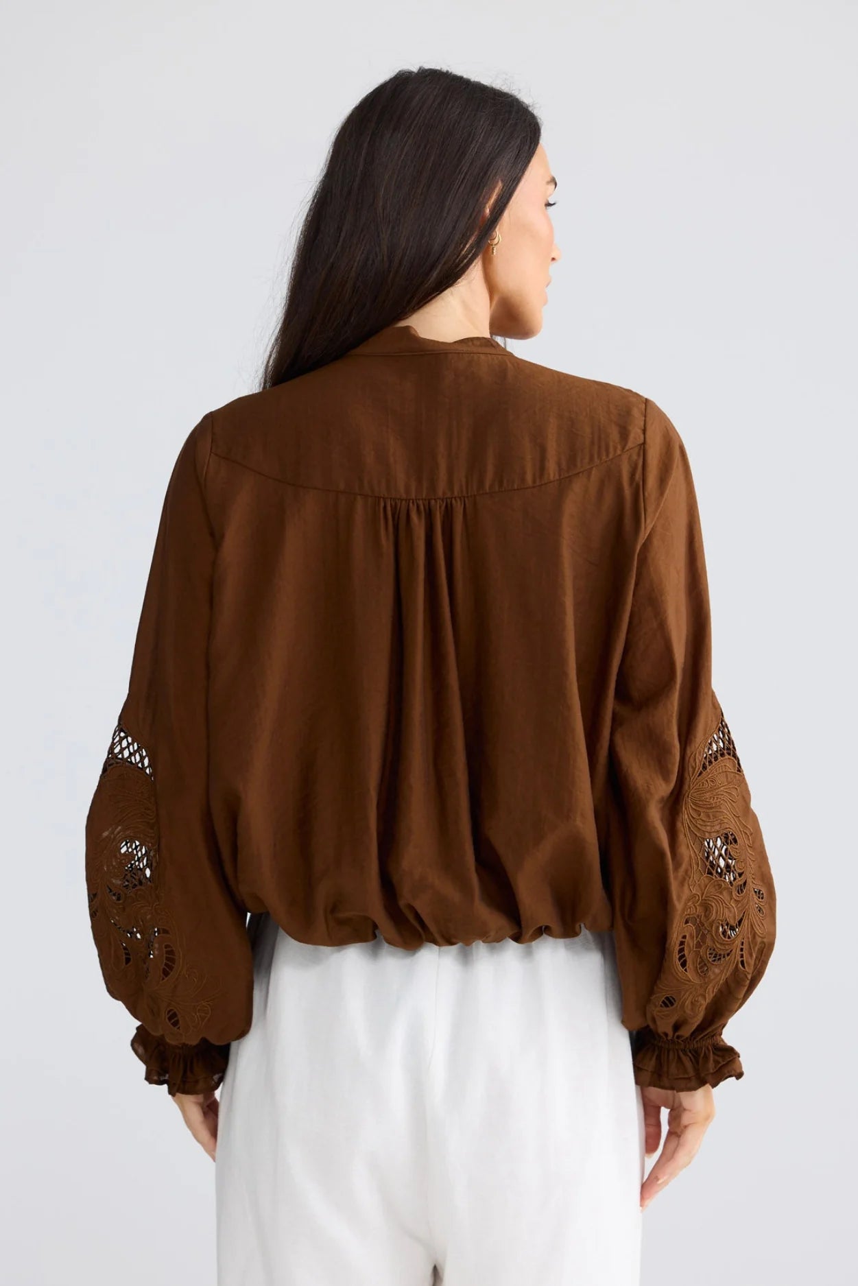 A woman viewed from the back wearing a brown long-sleeve blouse with embroidered details on the sleeves and white pants, set against a plain light gray studio background.