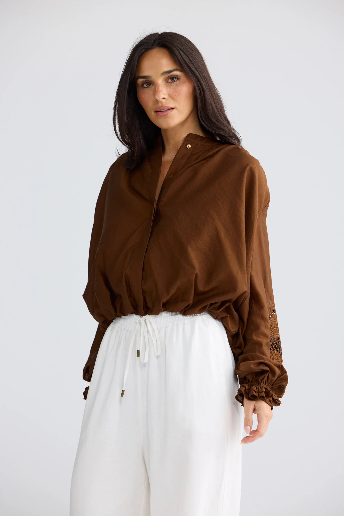 A woman modeling a brown long-sleeve button-up shirt with gathered cuffs, paired with white drawstring pants, against a light gray studio background.