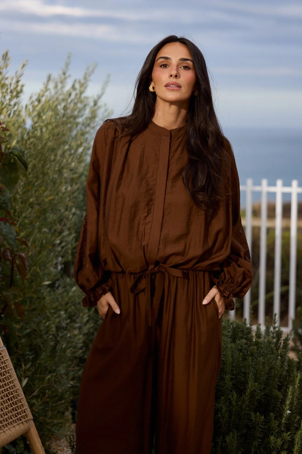 A woman wearing a brown long-sleeve jumpsuit with a tie waist and wide legs, standing outdoors in front of green plants and a white fence.