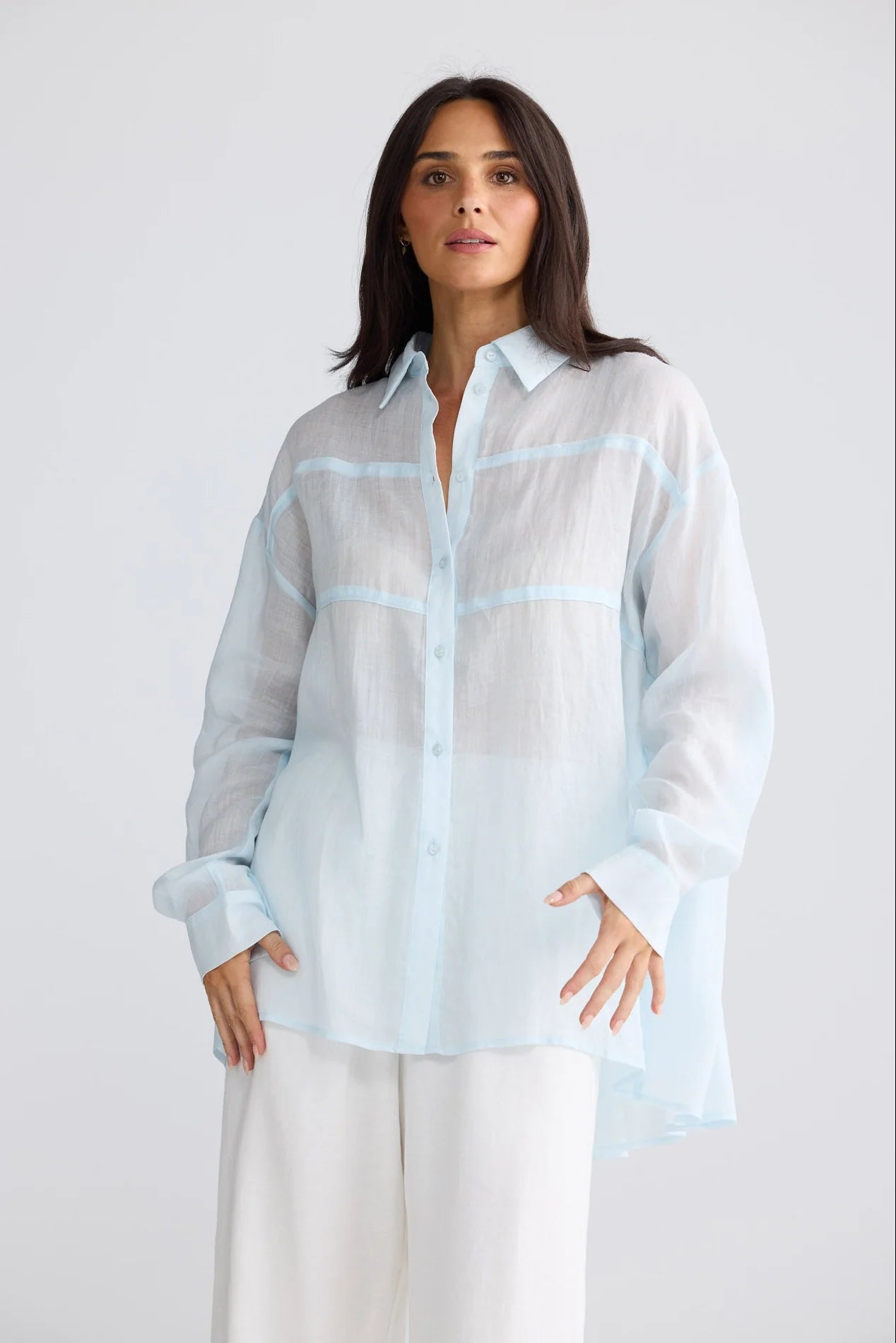 A woman modeling a light blue semi-sheer long-sleeve button-up shirt with white wide-leg pants, photographed against a clean light gray studio background.