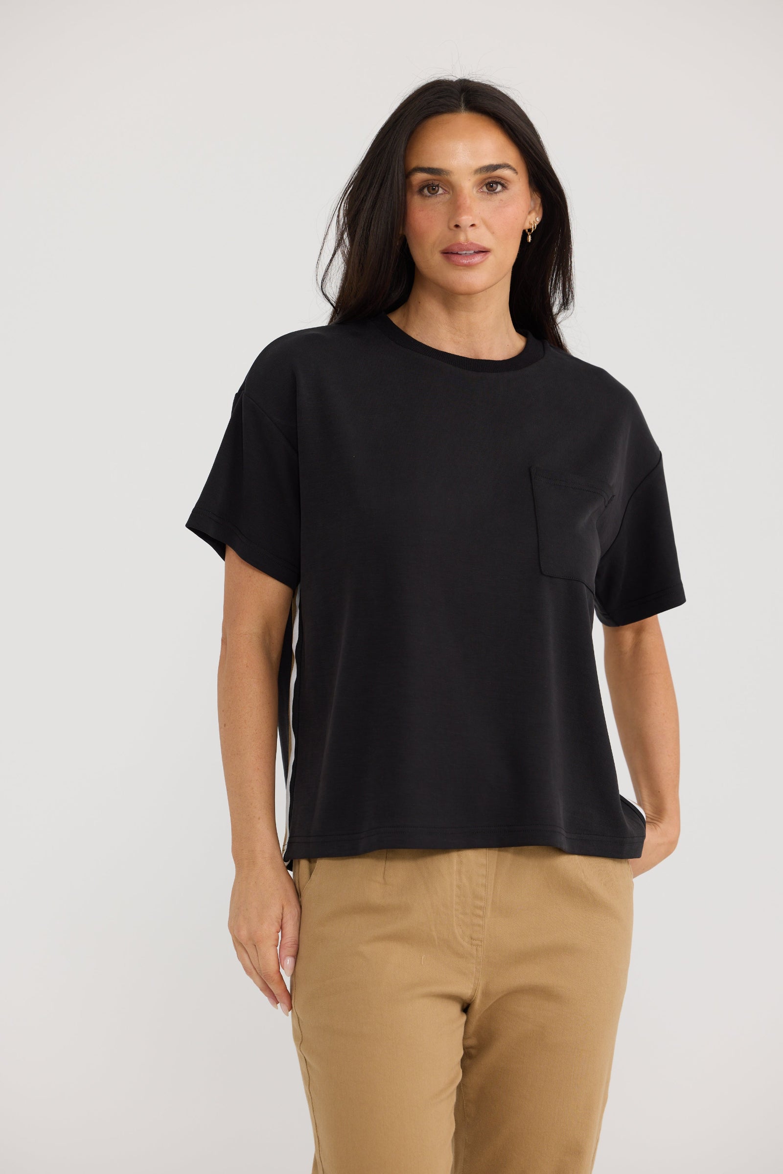 A woman modeling a black short-sleeve crewneck t-shirt with a chest pocket, paired with tan pants, against a plain light gray studio background.