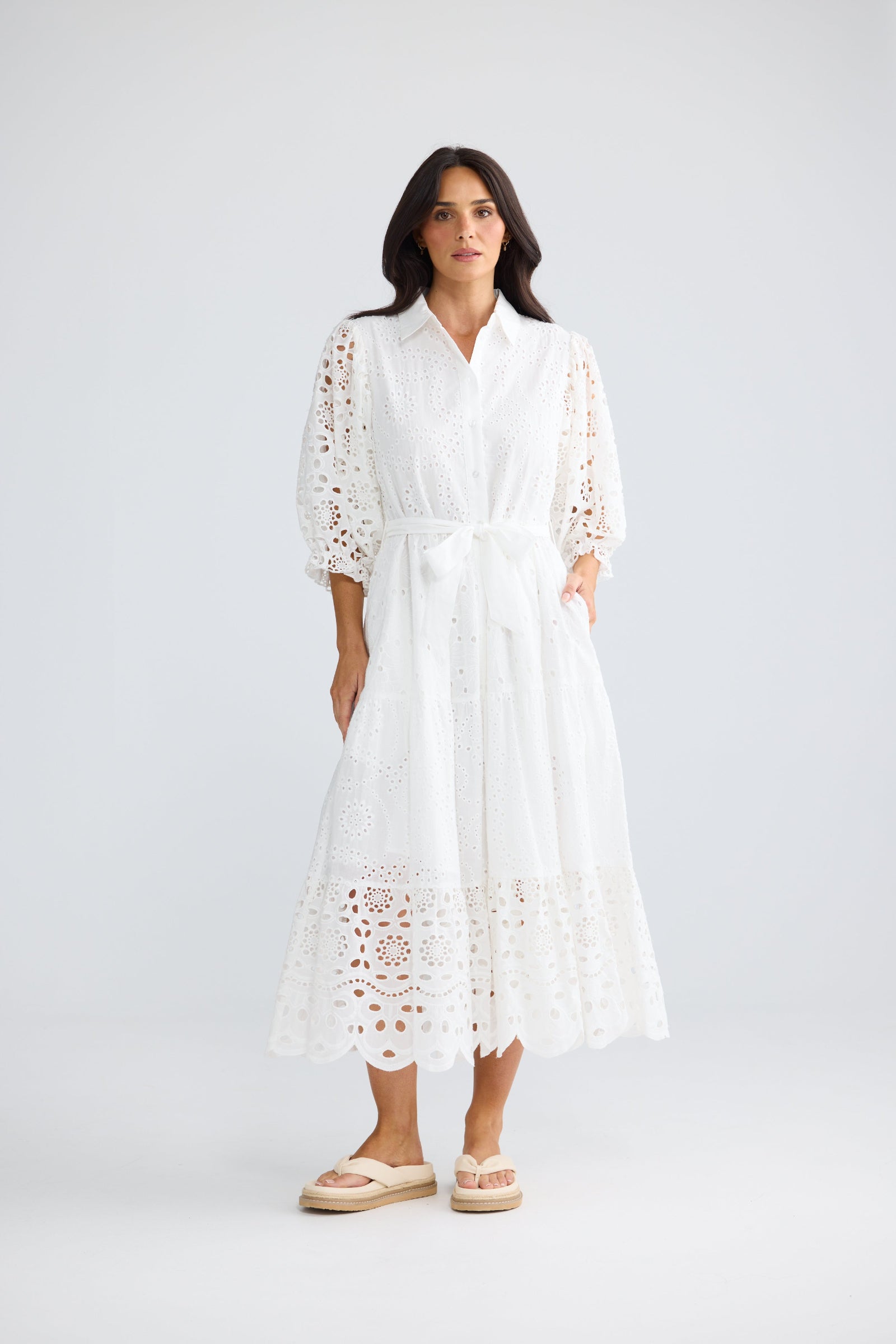 A woman modeling a white eyelet midi dress with three-quarter sleeves, a tie waist, and side pockets, paired with beige espadrille sandals against a light gray studio background.