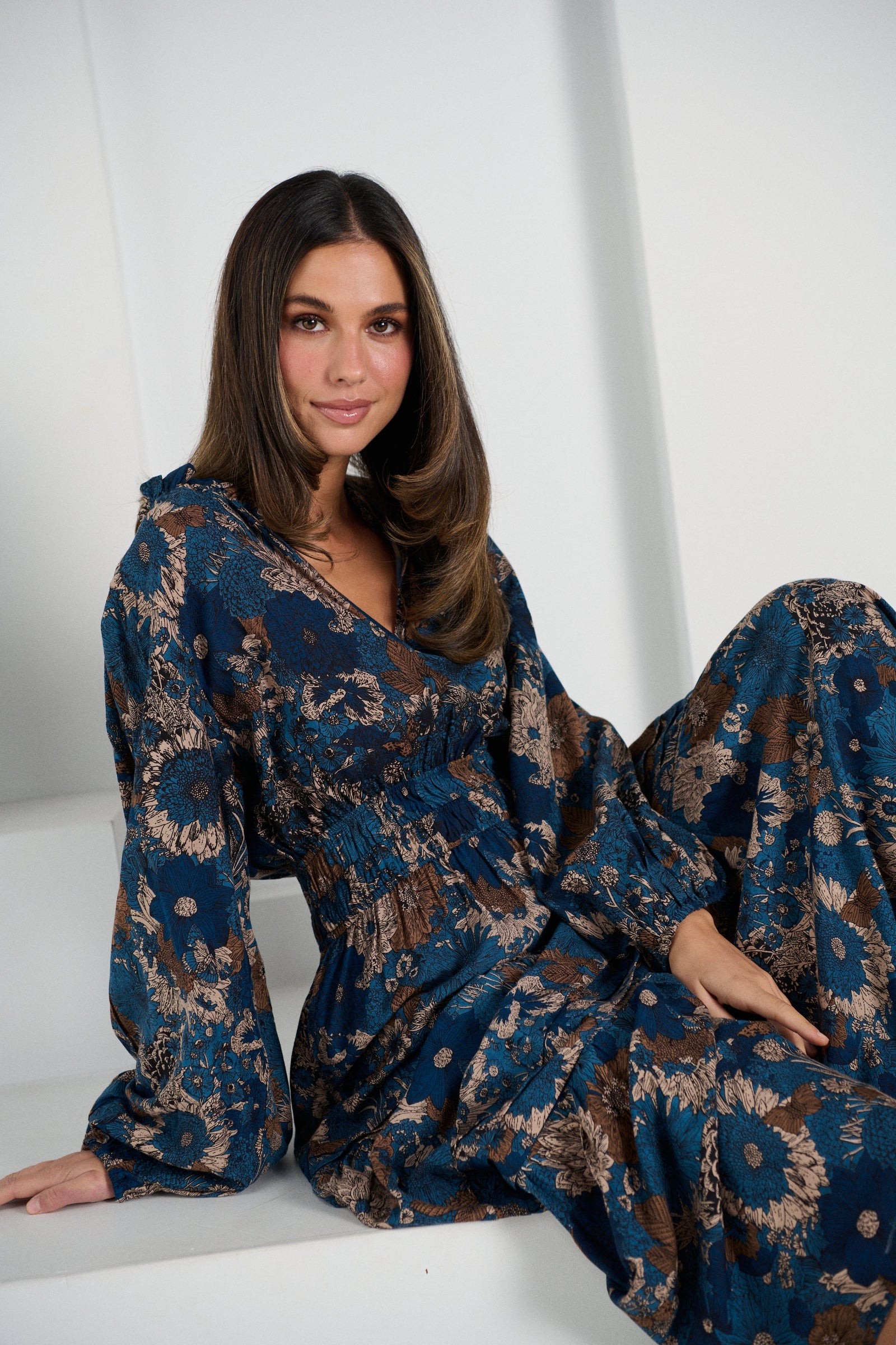 Woman wearing a blue floral dress sitting on a white surface with a white curtain background