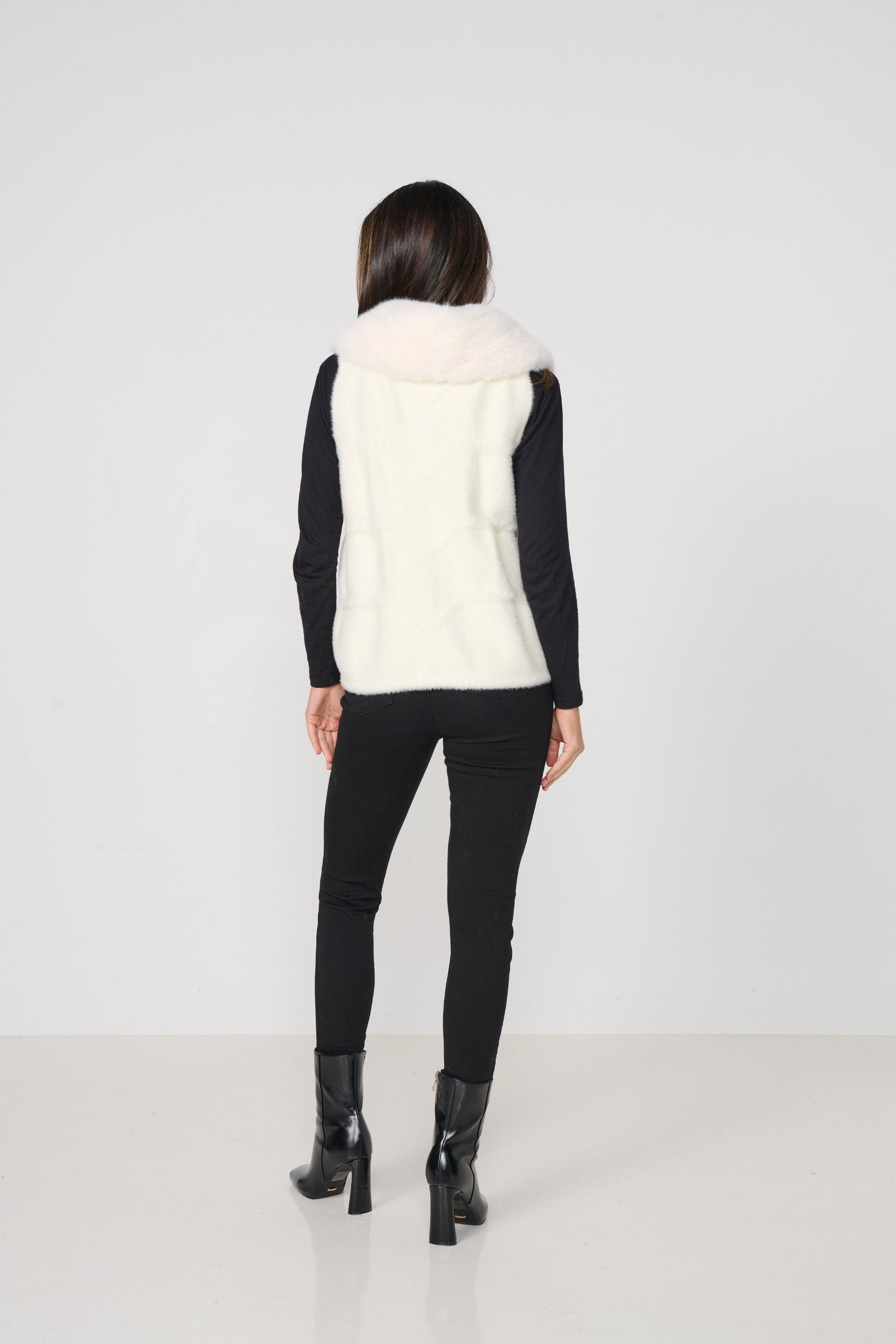 Person wearing a white fur vest over a black long-sleeve shirt and black pants on a white background