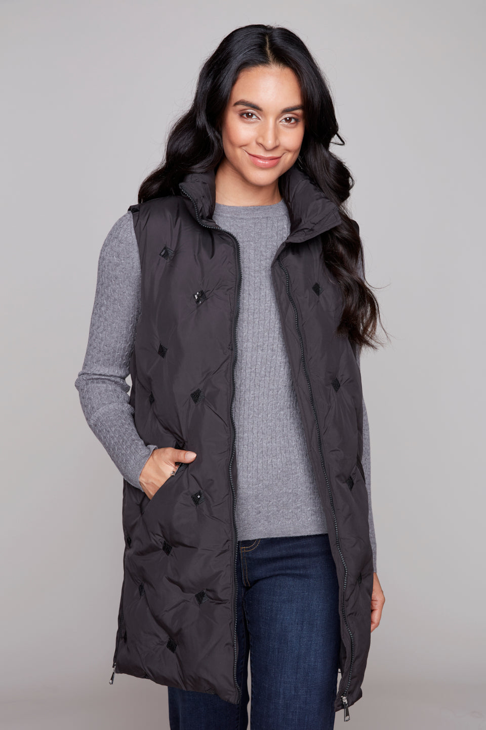 Woman wearing a black longline puffer vest over a gray sweater on a plain background