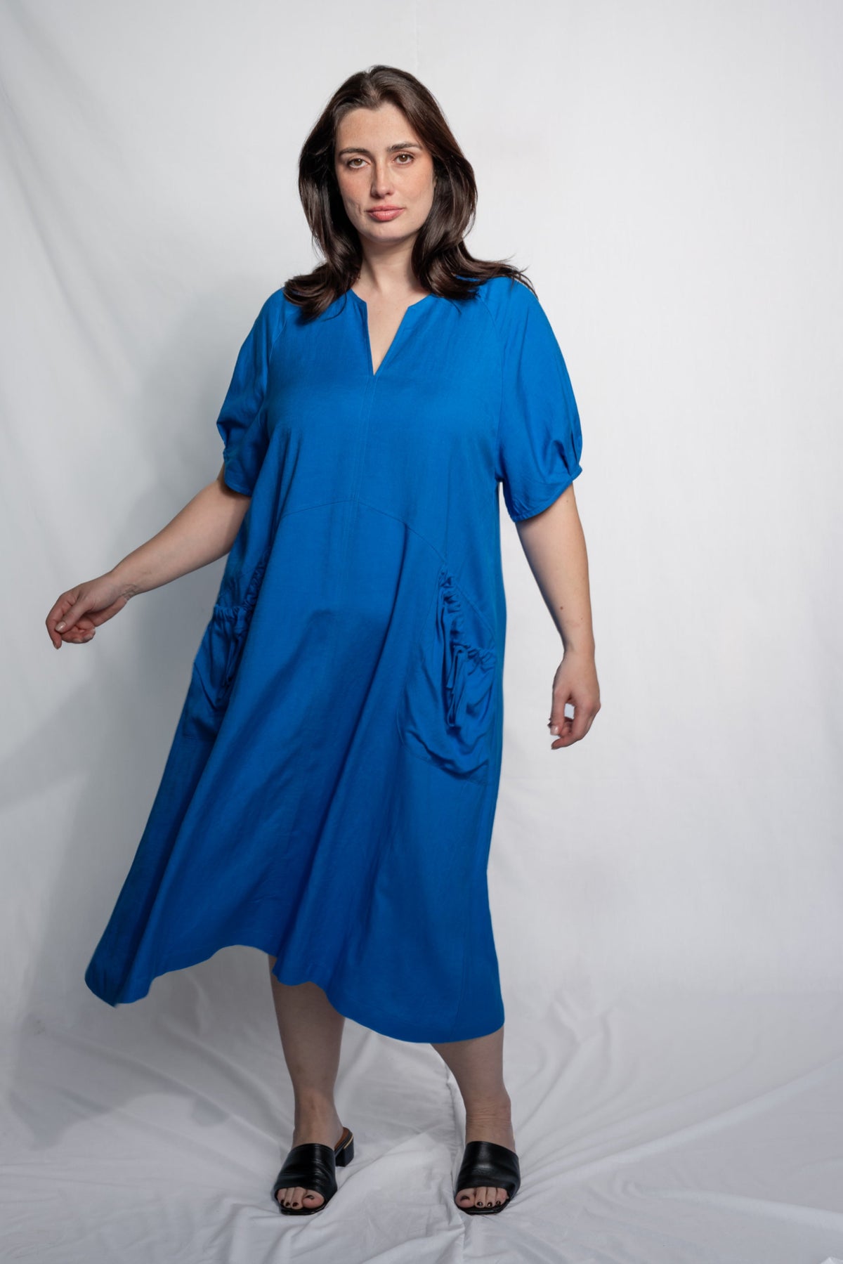 A woman modeling a bright blue midi dress with short puff sleeves, a split V-neckline, and side pockets, standing against a white studio backdrop.