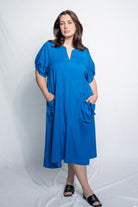A woman modeling a bright blue midi dress with short puff sleeves, a split neckline, and large patch pockets, standing against a white studio backdrop.