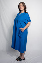 A woman modeling a bright blue midi dress with short sleeves, a split neckline, and a side pocket, standing against a white studio backdrop.