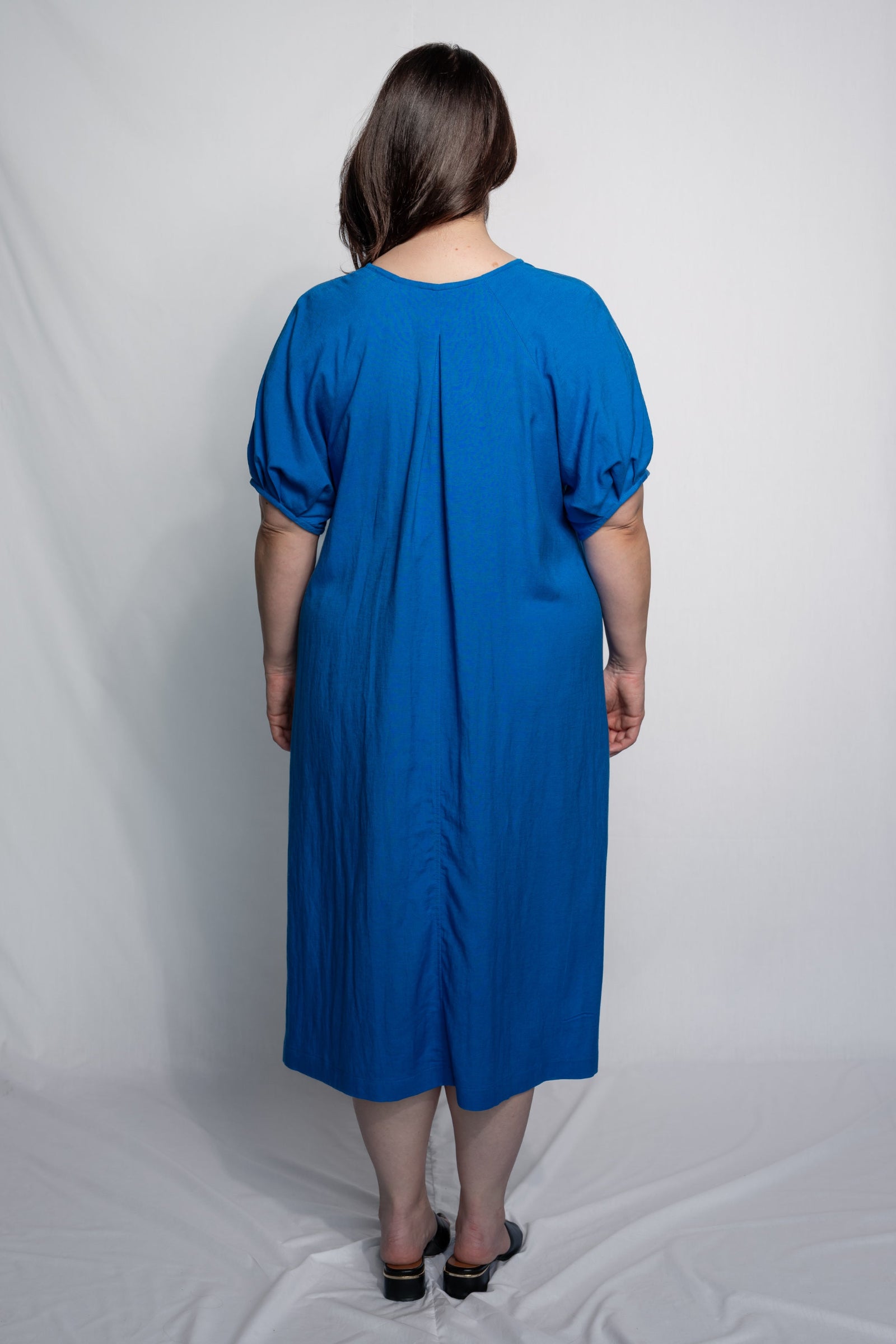 A woman viewed from behind wearing a bright blue midi dress with short puff sleeves and a gathered back seam, standing against a white studio backdrop.
