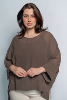 A woman modeling a loose-fitting, draped brown blouse with wide three-quarter sleeves and a round neckline, paired with light beige pants, against a light blue-gray studio background.