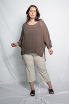 A woman modeling a loose, semi-sheer brown long-sleeve top paired with light beige cropped pants and black slide sandals, standing against a white draped studio backdrop.