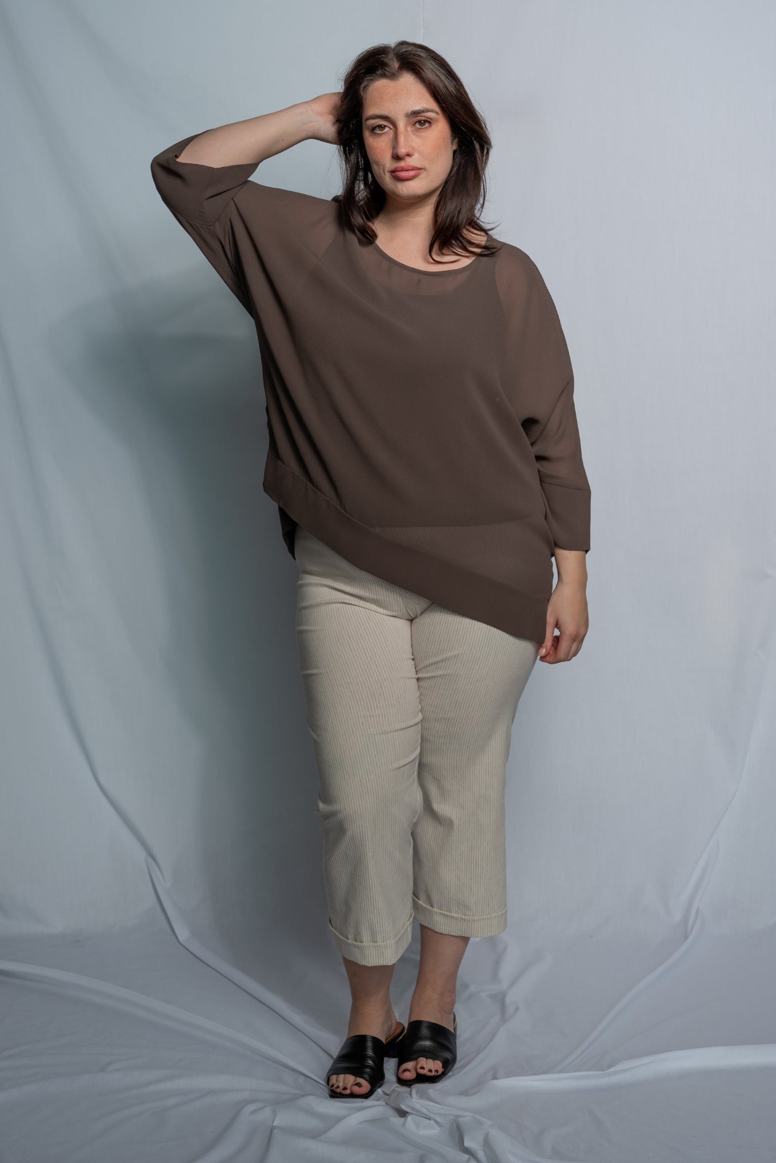A woman modeling a loose-fitting, three-quarter sleeve brown top paired with beige cropped pants and black slide sandals, standing against a light gray fabric backdrop.