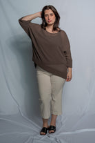 A woman modeling a loose-fitting, three-quarter sleeve brown top paired with beige cropped pants and black slide sandals, standing against a light gray fabric backdrop.