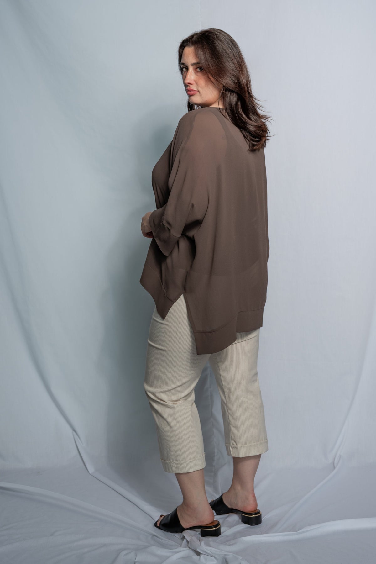 A woman modeling a loose, oversized taupe-brown top paired with beige cropped pants and black slide sandals, standing against a light gray studio backdrop.