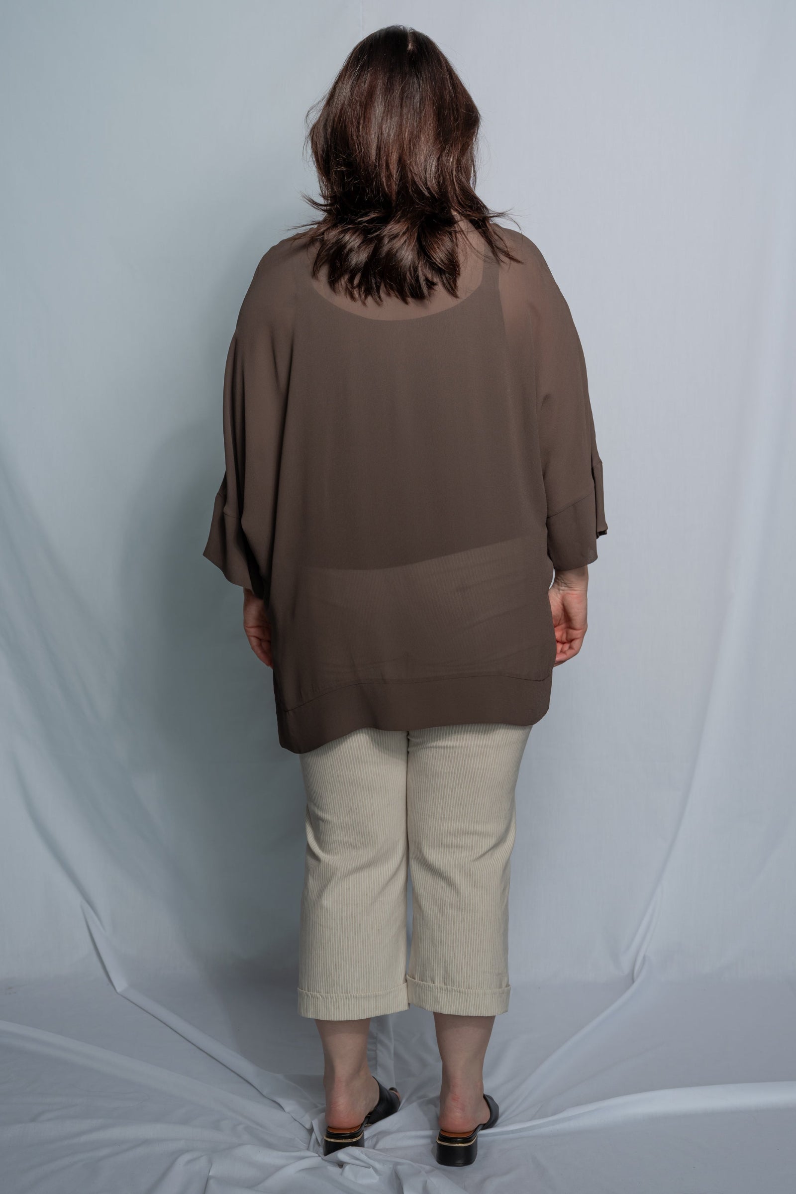 A woman viewed from behind wearing a loose, taupe-brown short-sleeve top and light beige cropped pants, standing against a draped light gray studio backdrop.
