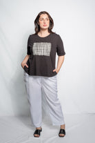 A woman modeling a black short-sleeve t-shirt with a white geometric grid pattern on the chest, paired with light gray wide-leg pants and black slide sandals, set against a light gray studio backdrop.