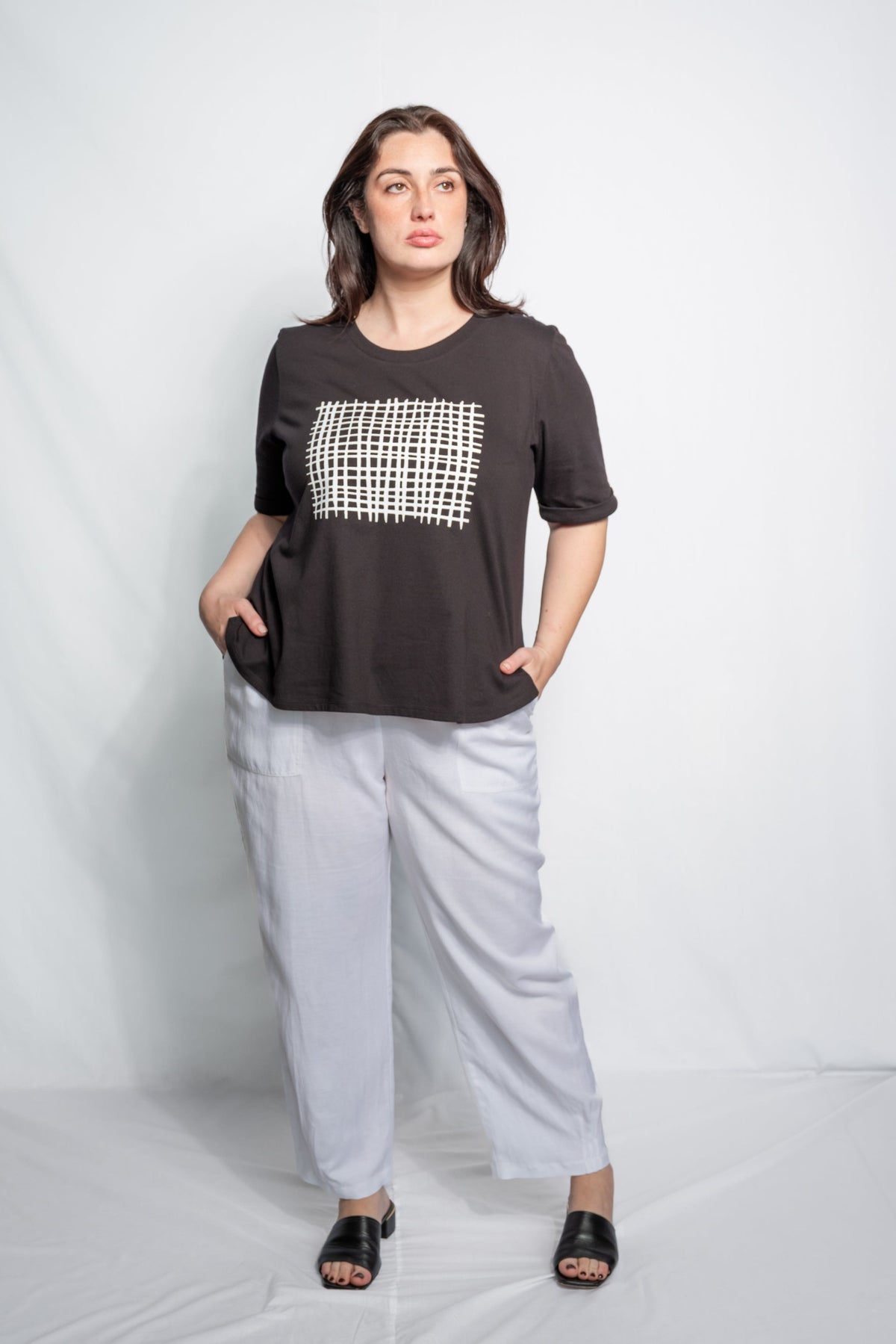 A woman modeling a black short-sleeve t-shirt with a white geometric grid pattern on the chest, paired with light gray wide-leg pants and black slide sandals, set against a light gray studio backdrop.