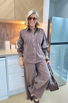 A woman wearing a matching brown corduroy button-up shirt and wide-leg pants set, holding a brown leather handbag in a modern kitchen setting.