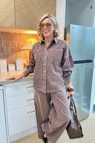 A woman wearing a matching brown corduroy button-up shirt and wide-leg trousers set, holding a brown leather bag in a modern kitchen setting.