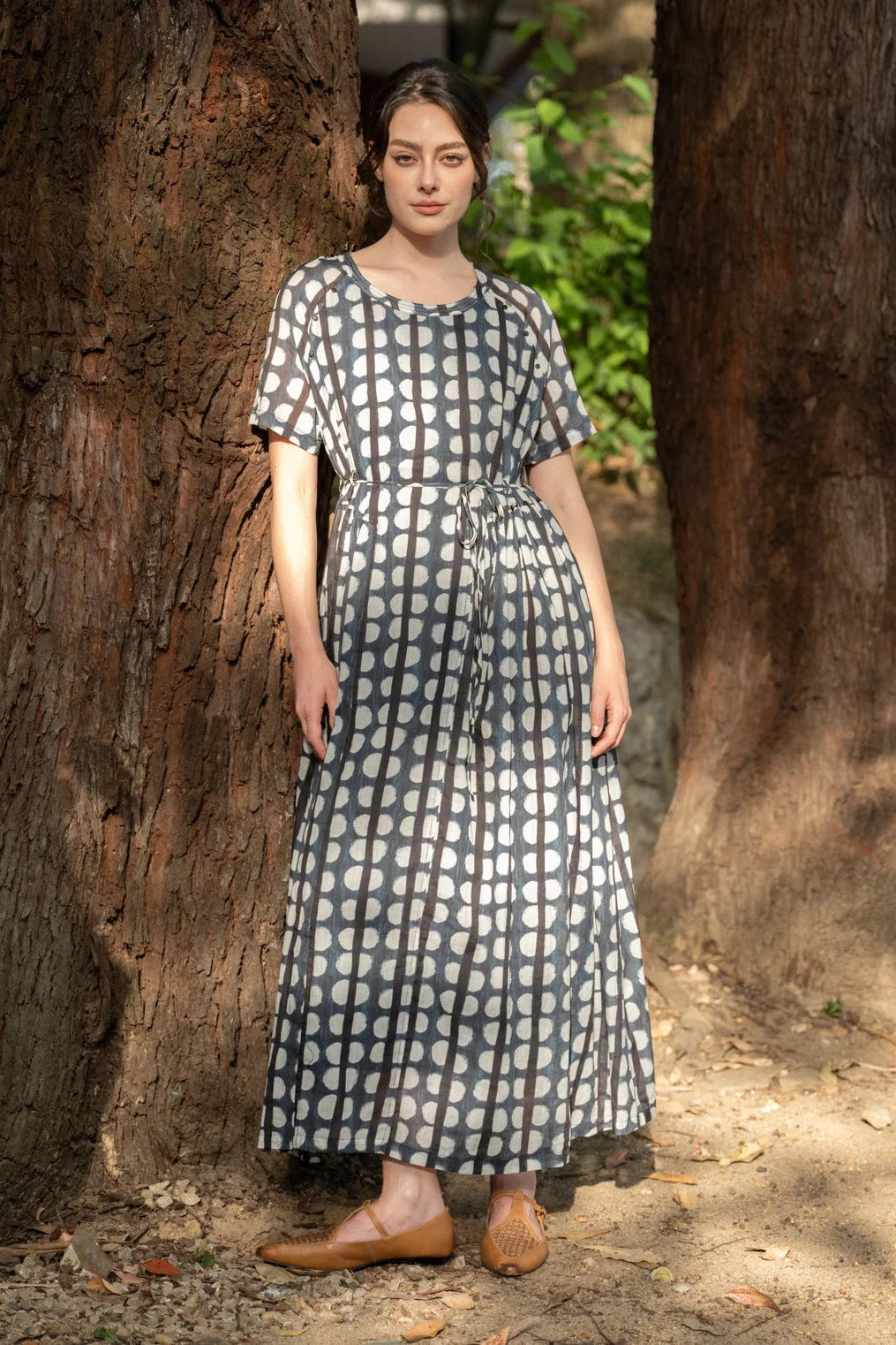 A woman wearing a short-sleeve midi dress with a white and gray polka dot pattern, standing outdoors between two large tree trunks.