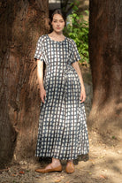 A woman wearing a short-sleeve midi dress with a white and gray polka dot pattern, standing outdoors between two large tree trunks.