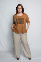 A woman modeling a brown short-sleeve top with a black grid pattern on the chest, paired with beige wide-leg trousers, standing against a light gray studio backdrop.