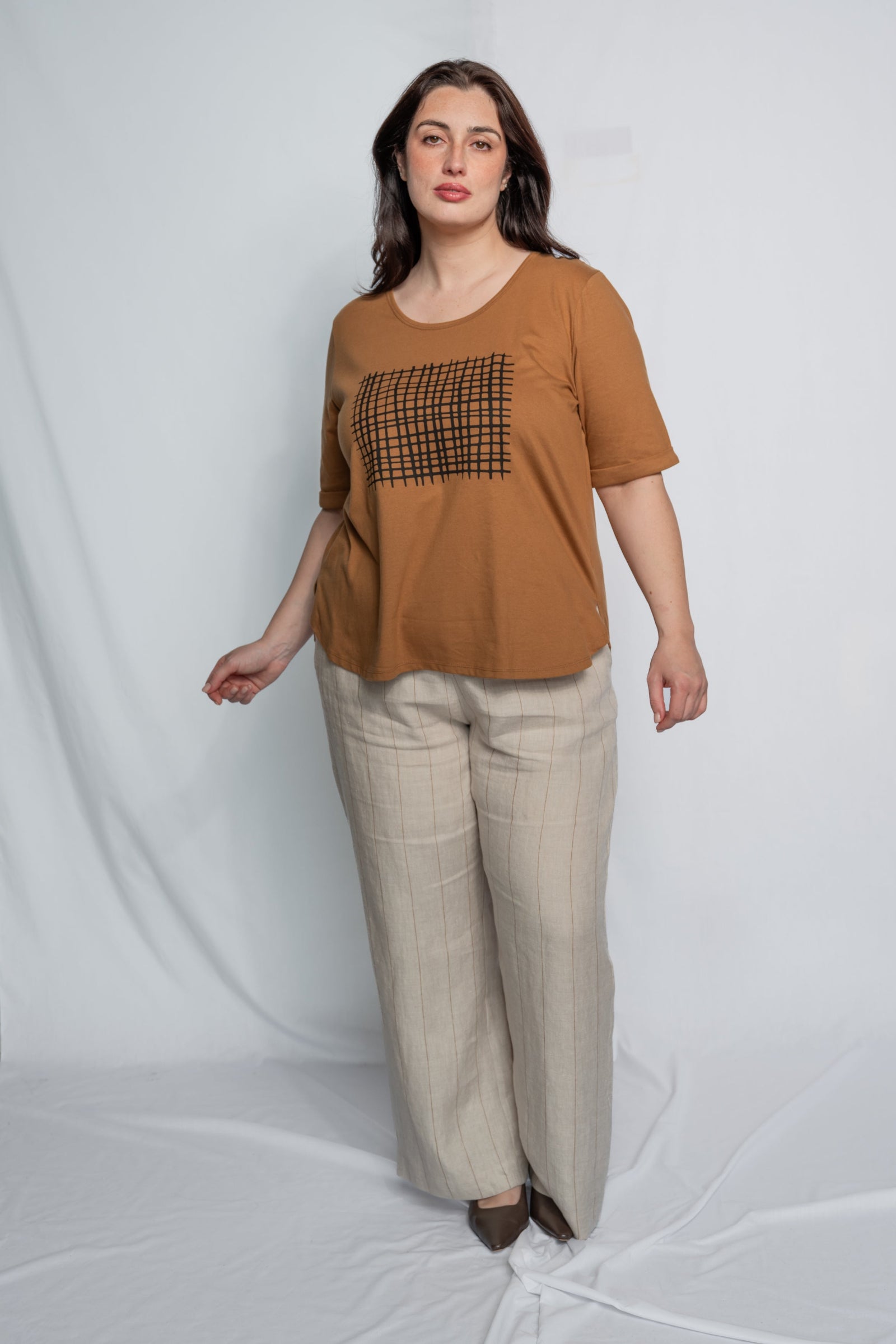 A woman modeling a brown short-sleeve t-shirt with a black checkered square graphic on the chest, paired with beige wide-leg trousers, standing against a white fabric backdrop.