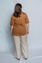 A woman viewed from behind wearing a rust-brown short-sleeve top and light beige wide-leg trousers, standing against a light blue studio backdrop.