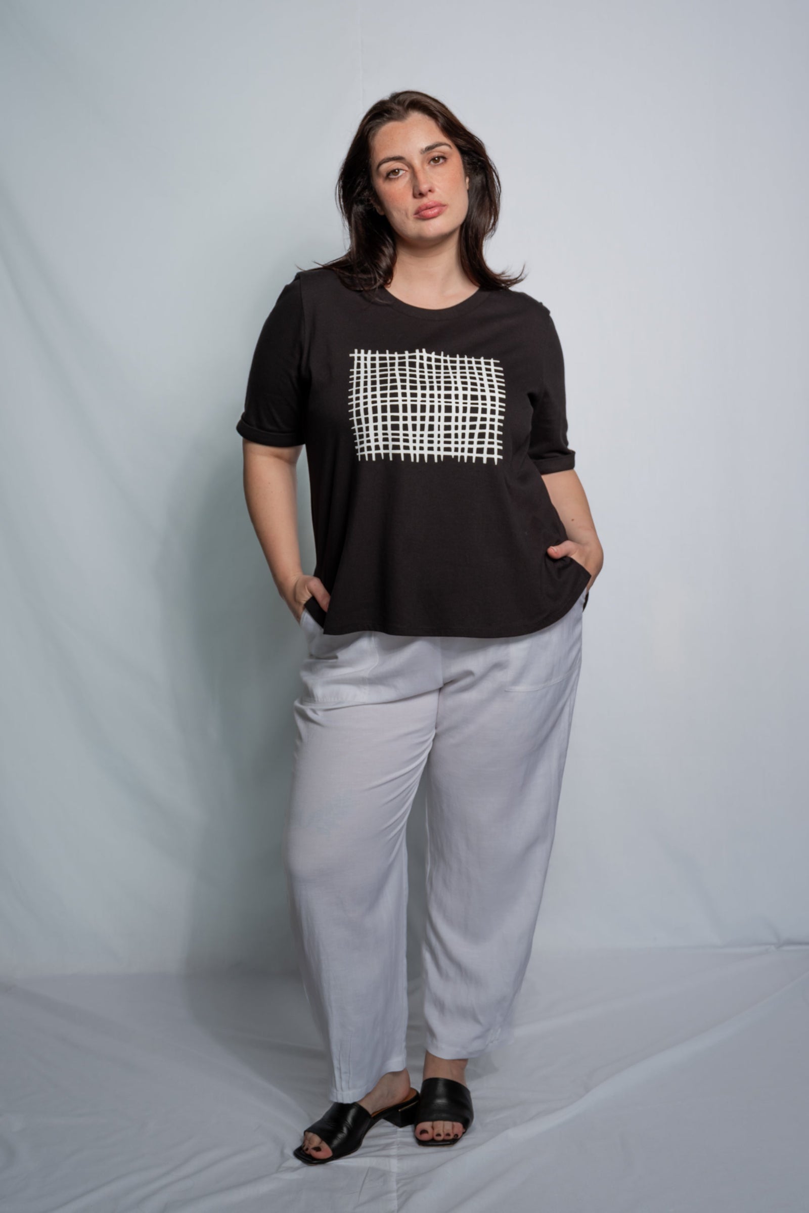 A woman modeling a black short-sleeve top with a white geometric patterned chest panel, paired with light gray wide-leg pants and black sandals, standing against a light gray studio backdrop.