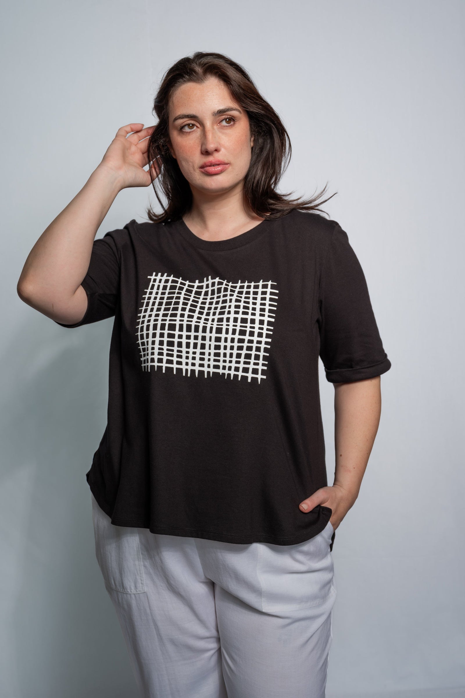 A woman modeling a black short-sleeve t-shirt with a white grid pattern graphic on the chest, paired with light gray pants, against a plain light gray background.