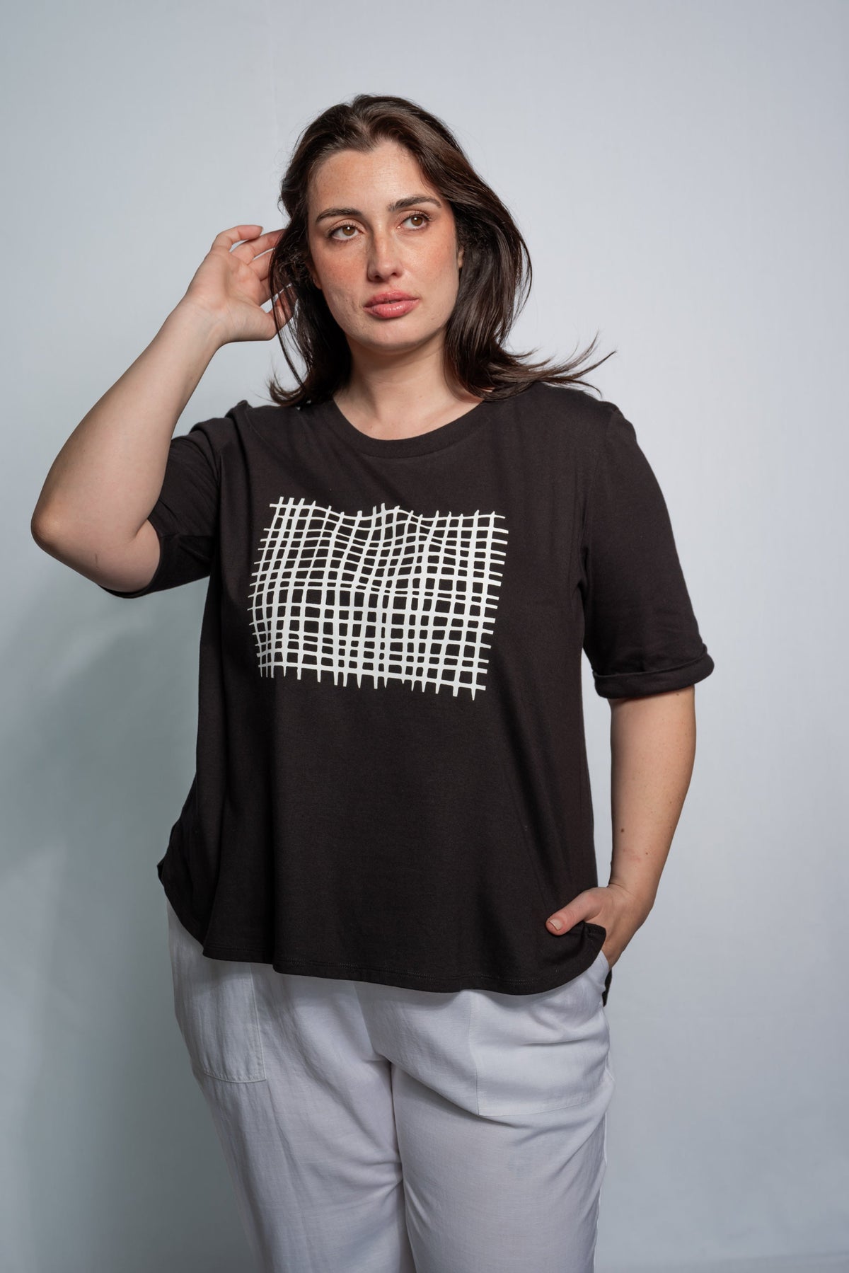 A woman modeling a black short-sleeve t-shirt with a white grid pattern graphic on the chest, paired with light gray pants, against a plain light gray background.