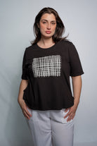 A woman modeling a black short-sleeve t-shirt with a white grid pattern printed on the chest, paired with light gray pants, against a plain light gray studio background.