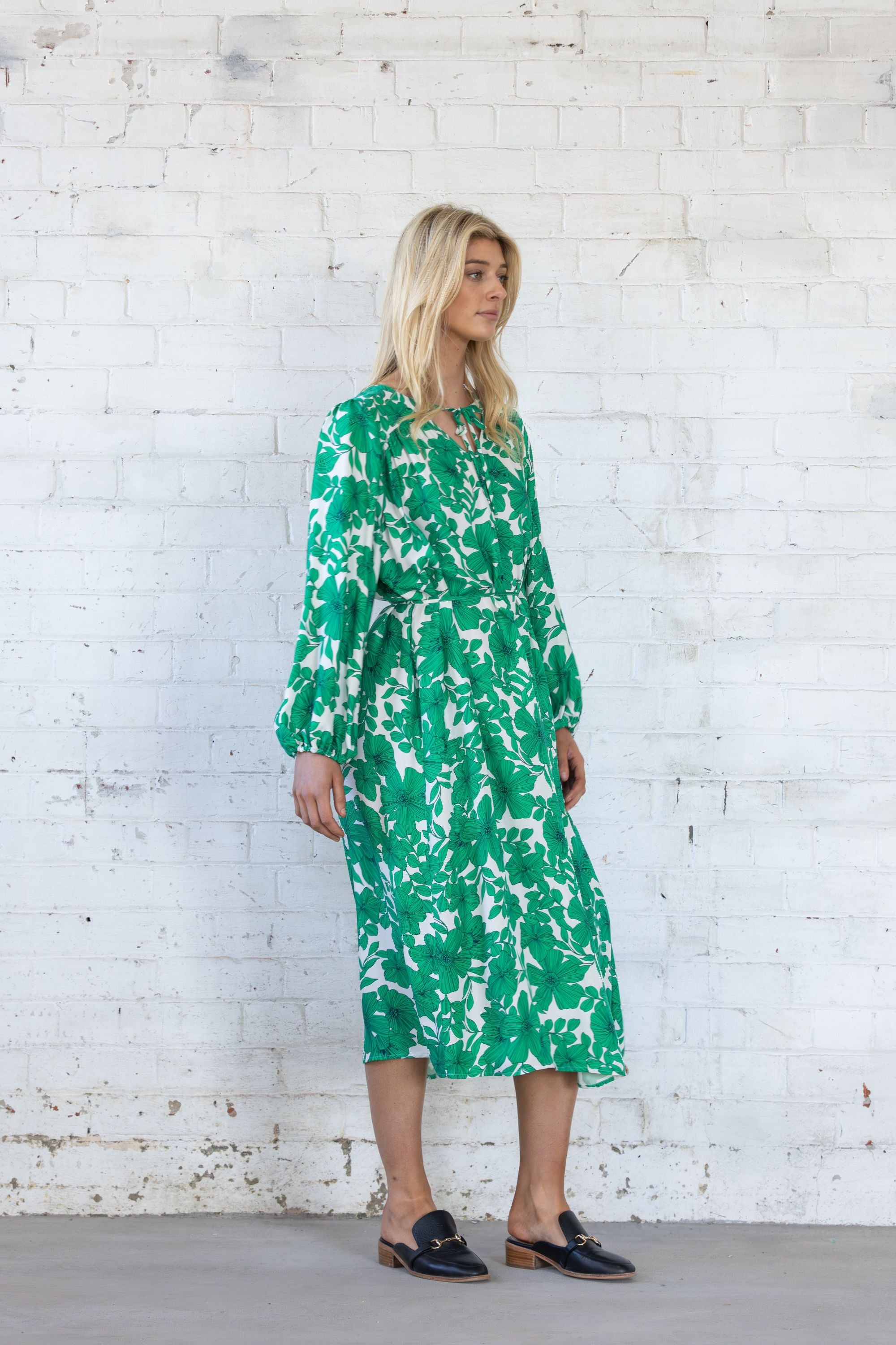 Woman wearing a green floral dress against a white brick wall.
