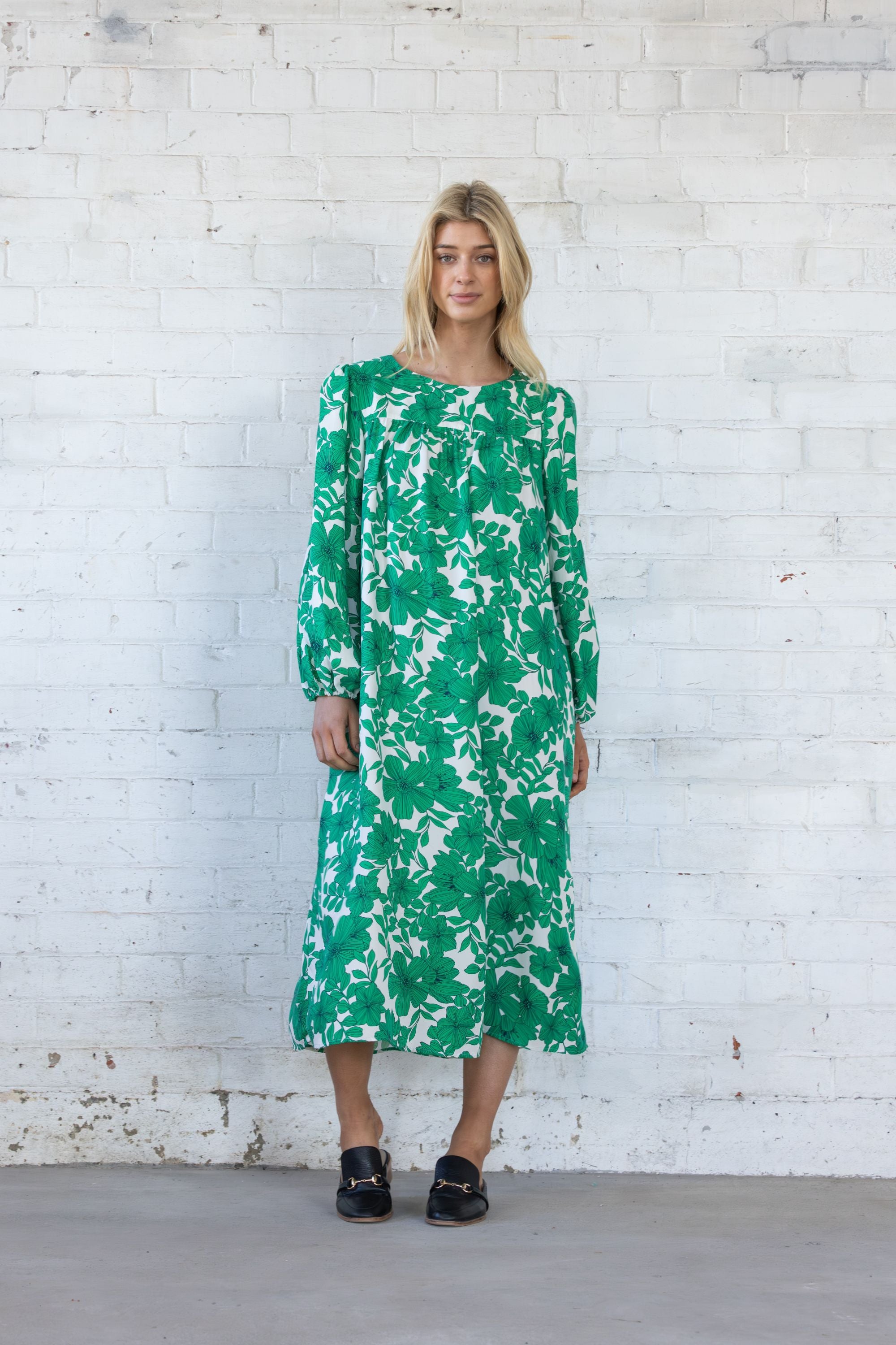 Woman wearing a green floral dress against a white brick wall.