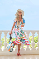 A woman stands on a white balcony wearing a colorful tropical print midi dress, a wide-brimmed straw hat, and red heels, while holding a matching tote bag.