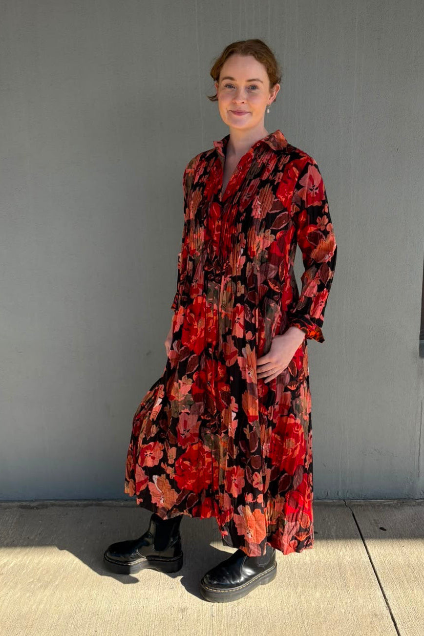 A woman wearing a long-sleeved, button-front midi dress with a bold red, orange, and black floral pattern, paired with black boots, standing outdoors against a gray concrete wall.