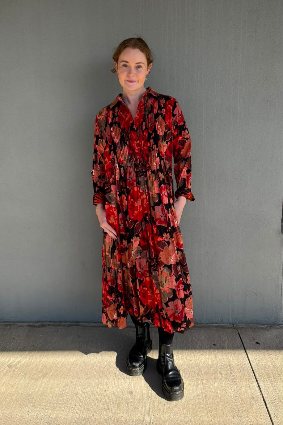 A woman stands outdoors against a gray building wall wearing a long-sleeve midi shirt dress with a bold red, black, and orange floral pattern, paired with black boots.