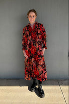 A woman stands outdoors against a gray building wall wearing a long-sleeve midi shirt dress with a bold red, black, and orange floral pattern, paired with black boots.